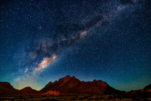 A sweeping view of the night sky above the Namib Desert, featuring the Milky Way galaxy glowing brightly in a diagonal arc. The foreground shows rugged desert mountains in silhouette, under a sky densely packed with stars.