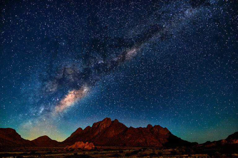 A sweeping view of the night sky above the Namib Desert, featuring the Milky Way galaxy glowing brightly in a diagonal arc. The foreground shows rugged desert mountains in silhouette, under a sky densely packed with stars.