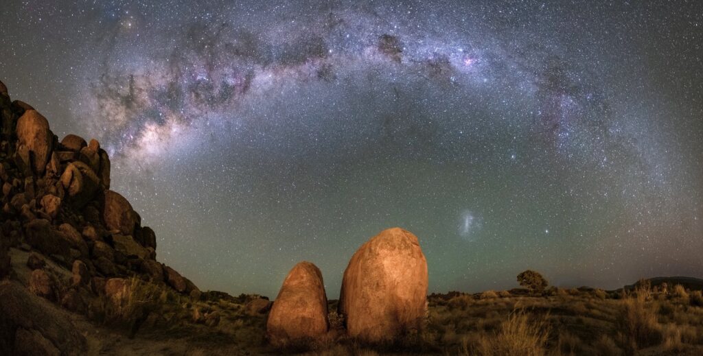 A luminous Milky Way arcs across the Namib Desert night sky. Large boulders and rocky outcrops dominate the foreground, softly lit under the starlight. Countless stars fill the sky in sharp detail, with a hint of green airglow near the horizon and the faint Magellanic Cloud visible.