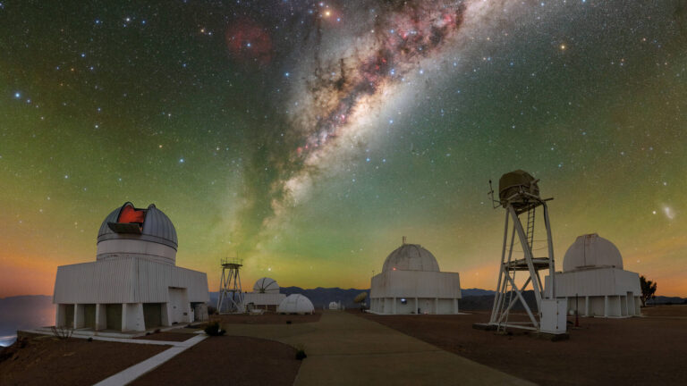 A row of observatory domes under the glowing arc of the Milky Way in the Atacama Desert at night. The sky is densely packed with stars and cosmic dust, with a green airglow near the horizon. Scientific instruments and antennas are silhouetted against the illuminated nightscape.
