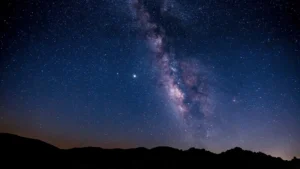 The Milky Way stretches vertically through a clear, star-filled sky above the dark silhouette of desert hills. A dense cluster of stars forms the galactic core, glowing in pink, white, and lavender hues. The foreground remains in deep shadow, enhancing the contrast of the sky.