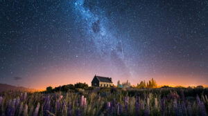 A small stone church sits under a sky bursting with stars and a glowing band of the Milky Way. A meteor streaks downward through the galactic plane. In the foreground, blooming lupines are visible in soft light, and a warm horizon glow suggests distant town lights.