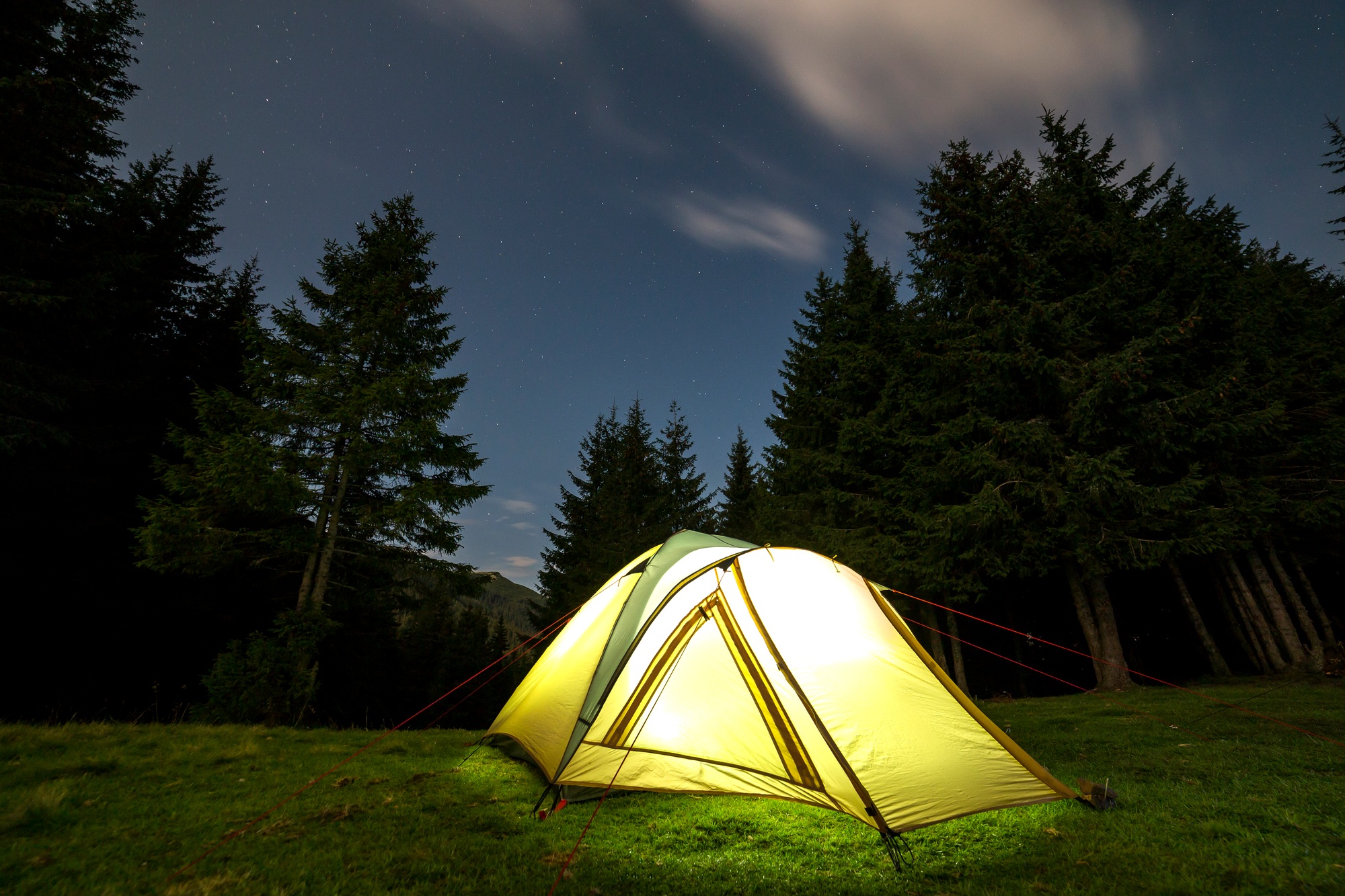 Summer camping at night. Illuminated tourist tent on green clearing on distant mountain background.